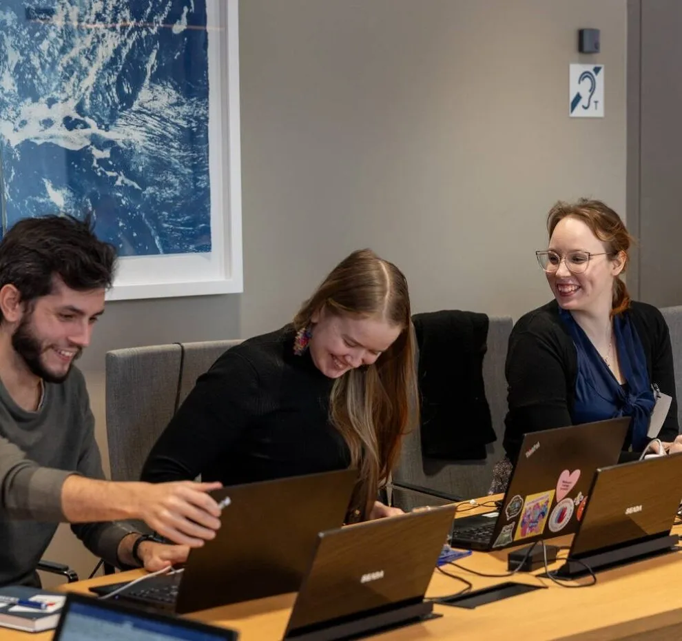 Three young people sitting in a meeting room with laptops, smiling at each other.