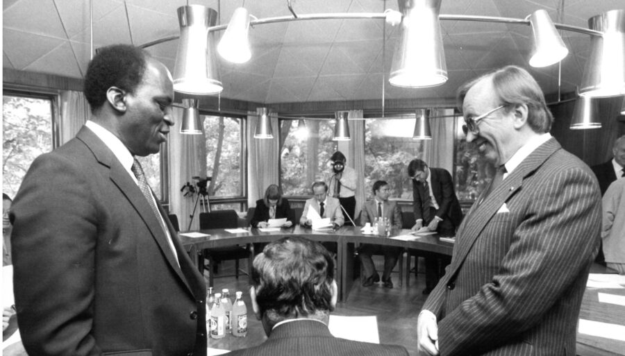 Two men in the foreground looking at each other, with people seated at a table in the background.