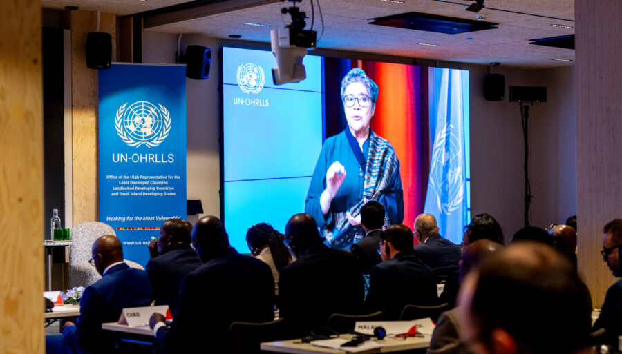 An image from an event showing an audience and a large screen with a speaker appearing on it.