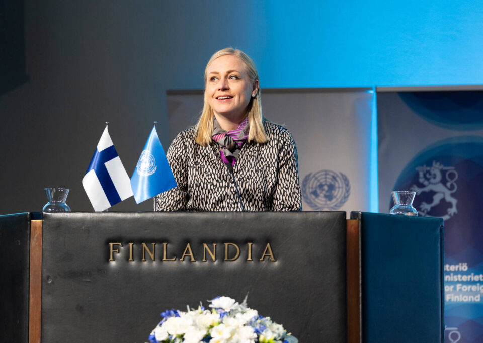 Minister for Foreign Affairs Elina Valtonen standing behind a podium and speaking to an audience, with the UN and Finnish flags in front.