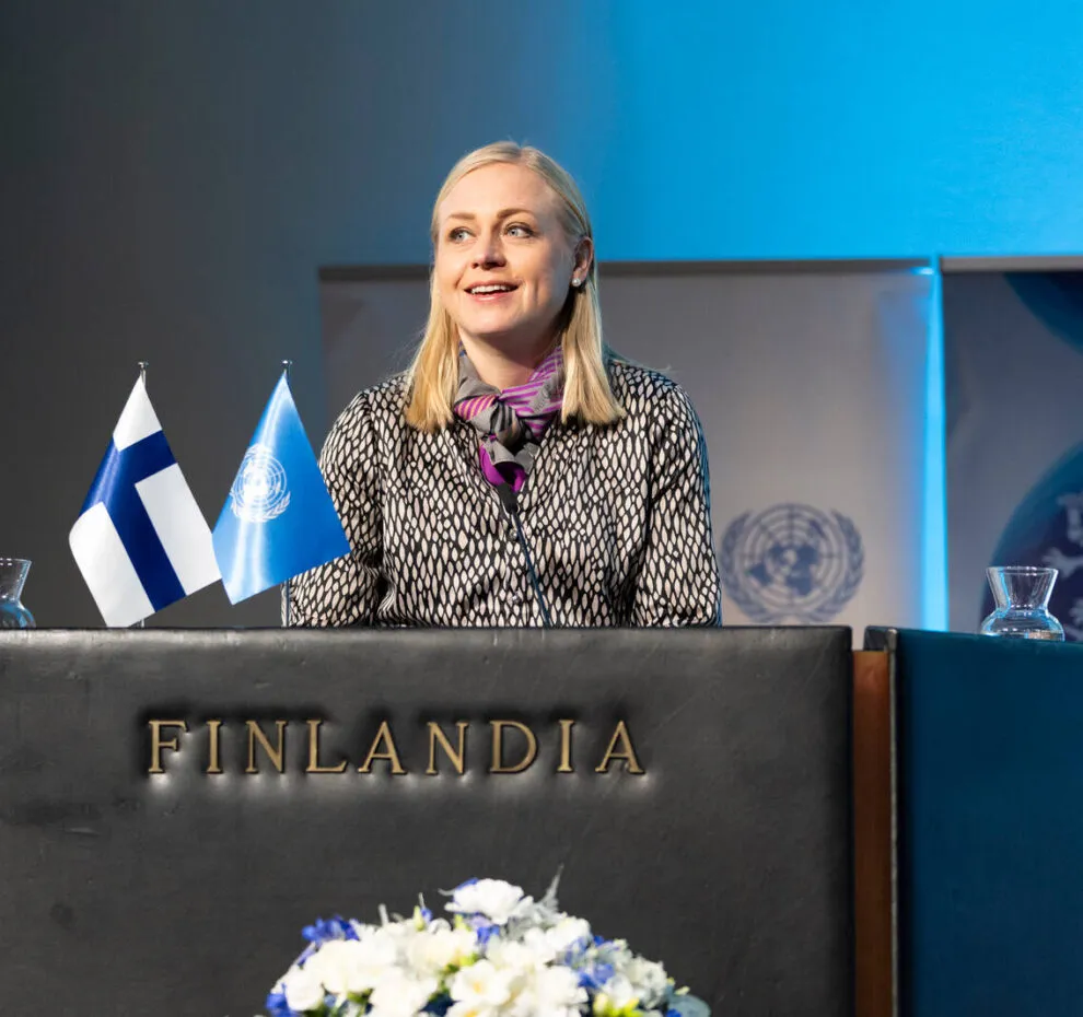 Minister for Foreign Affairs Elina Valtonen standing behind a podium and speaking to an audience, with the UN and Finnish flags in front.