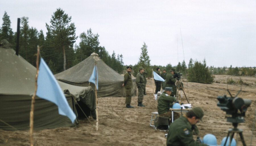 Military observers training in a shooting range area, with tents in the background.