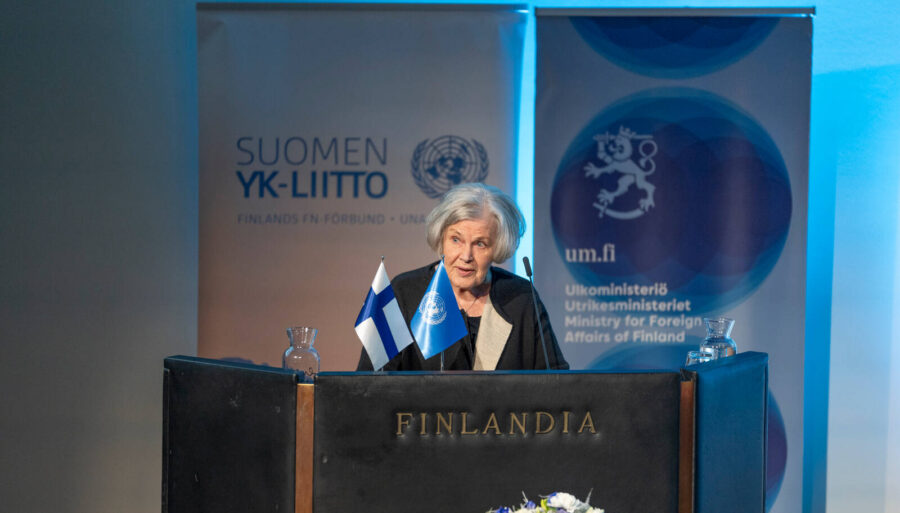 Former Defence Minister Elisabeth Rehn addressing the audience from behind the podium, with Finnish and UN flags in front of her.