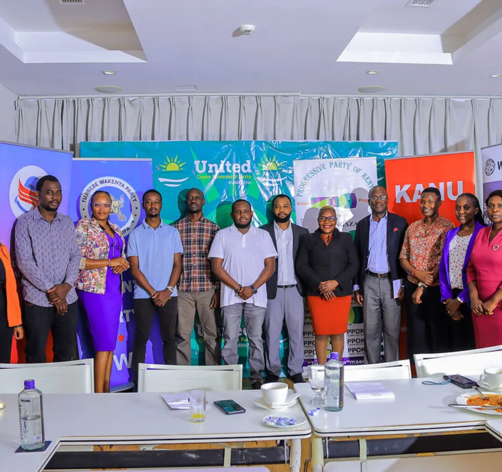 A group of people in a meeting room in Kenya, looking at the camera, with banners representing different political parties in the background.