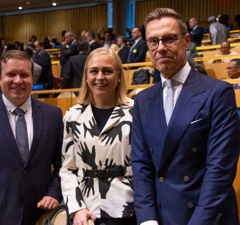 Minister Ville Tavio, Minister Elina Valtonen, and president of the republic Alexander Stubb standing and smiling behind Finland's place at the UN General Assembly hall.